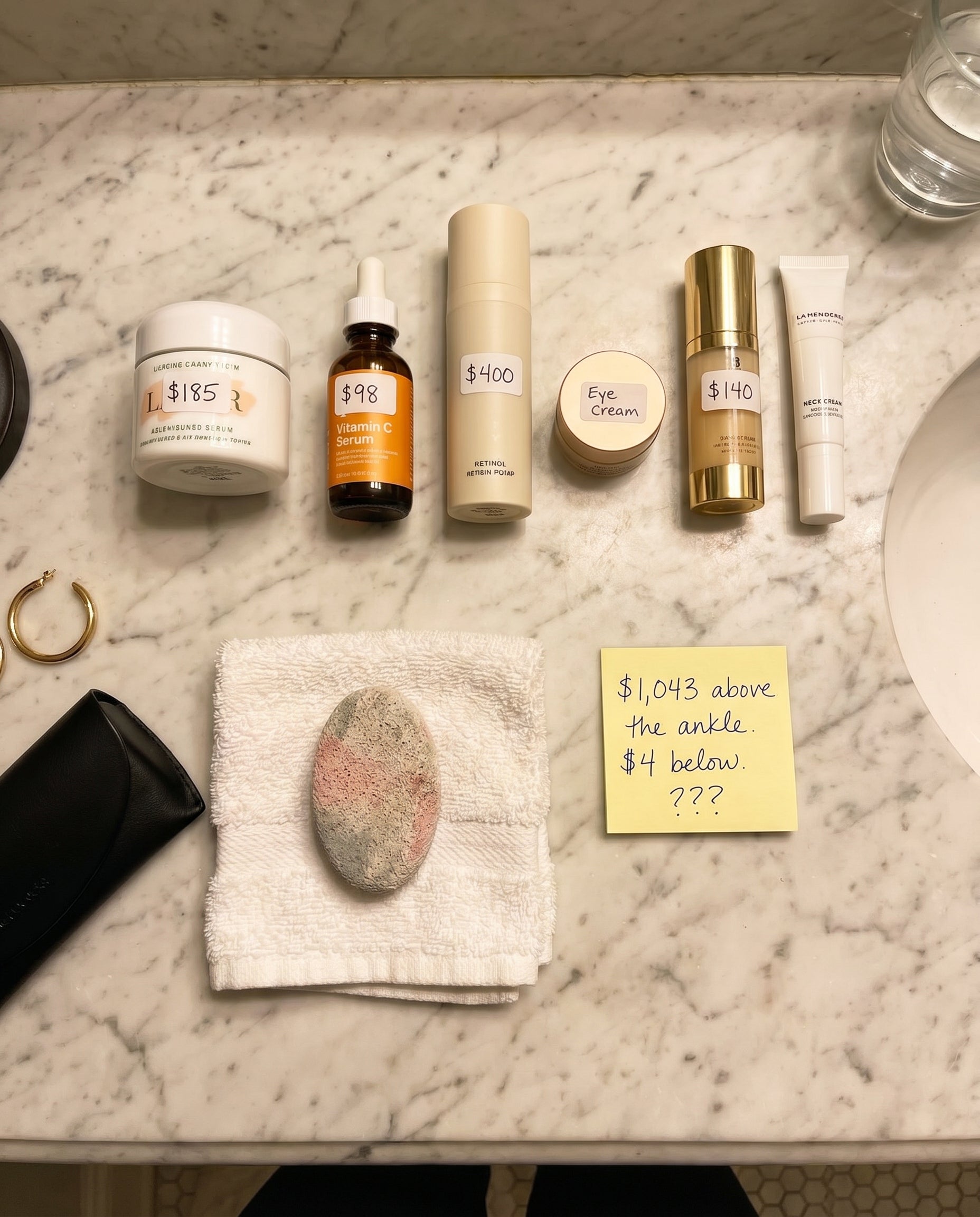 Overhead photo of a bathroom vanity showing several luxury skincare products with prices labeled, a small pumice stone on a washcloth, and a yellow sticky note reading $1,043 above the ankle, $4 below